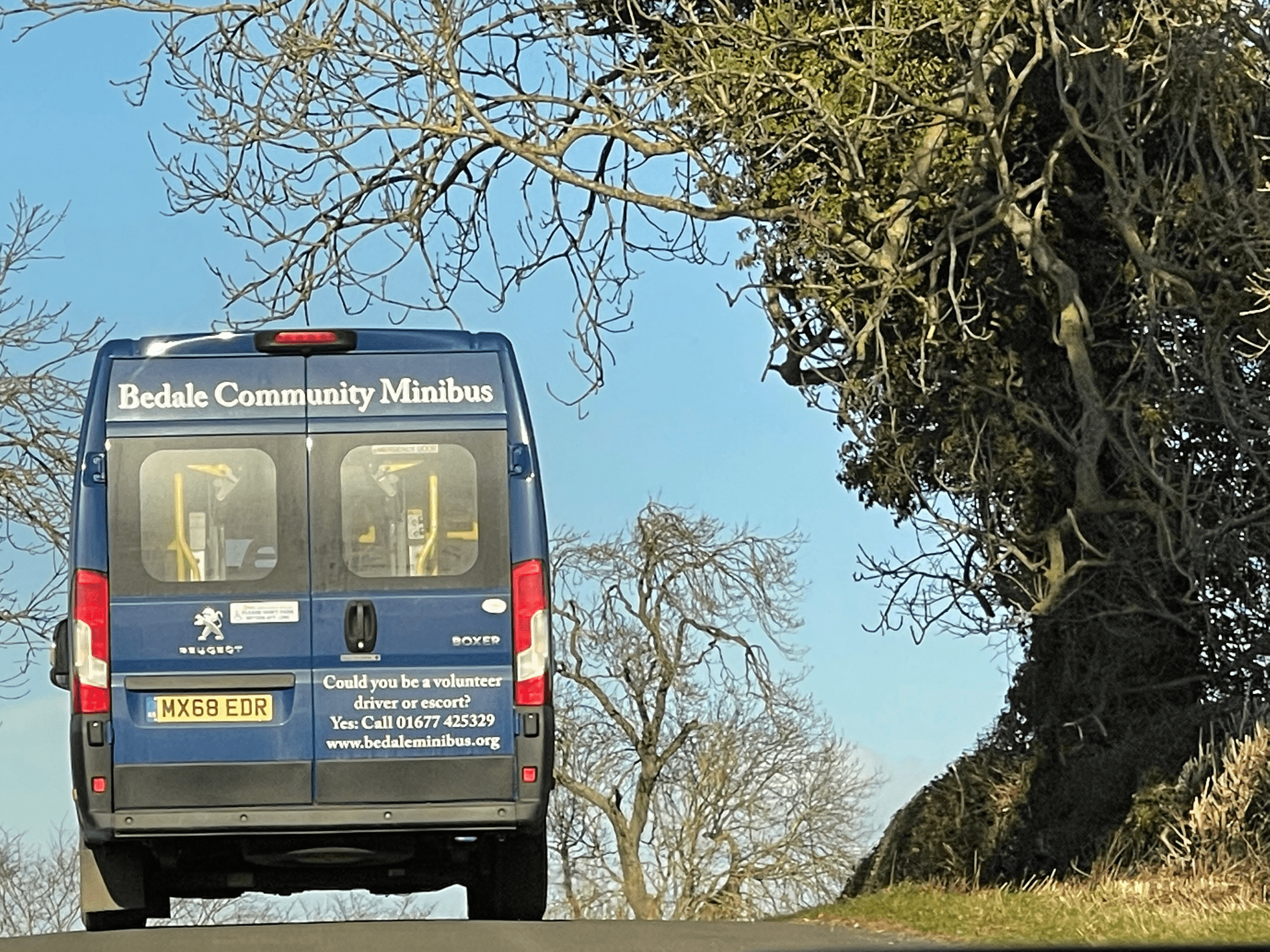 A Bedale Community Minibus Bus Traveling down a Tree Lined Back Road